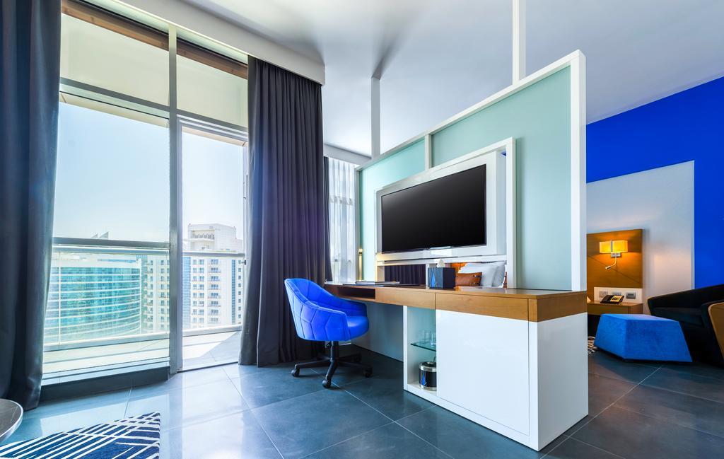 Hotel Tryp By Wyndham Dubái Exterior foto The photo shows a modern room with large windows allowing natural light to enter. There's a stylish desk area featuring a flat-screen TV mounted on the wall, along with a light wooden desk and a blue chair. The wall behind the TV is painted in a ligh